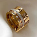 A gold-tone ring with Roman numerals and a central row of clear stones, photographed on a soft white fabric background.
