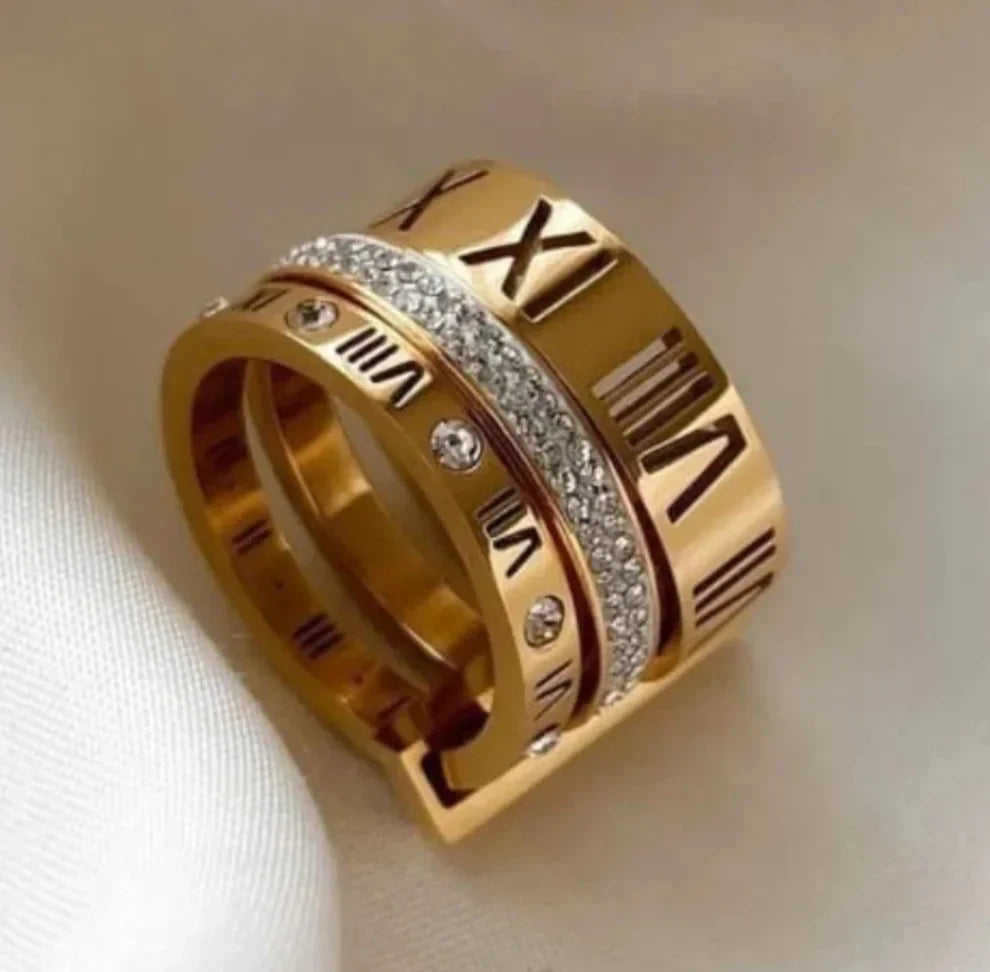 A gold-tone ring with Roman numerals and a central row of clear stones, photographed on a soft white fabric background.