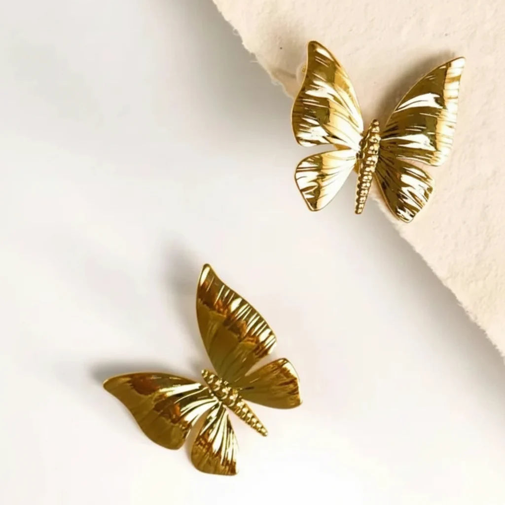Two gold butterfly-shaped earrings displayed on a white surface with a beige textured paper accent.