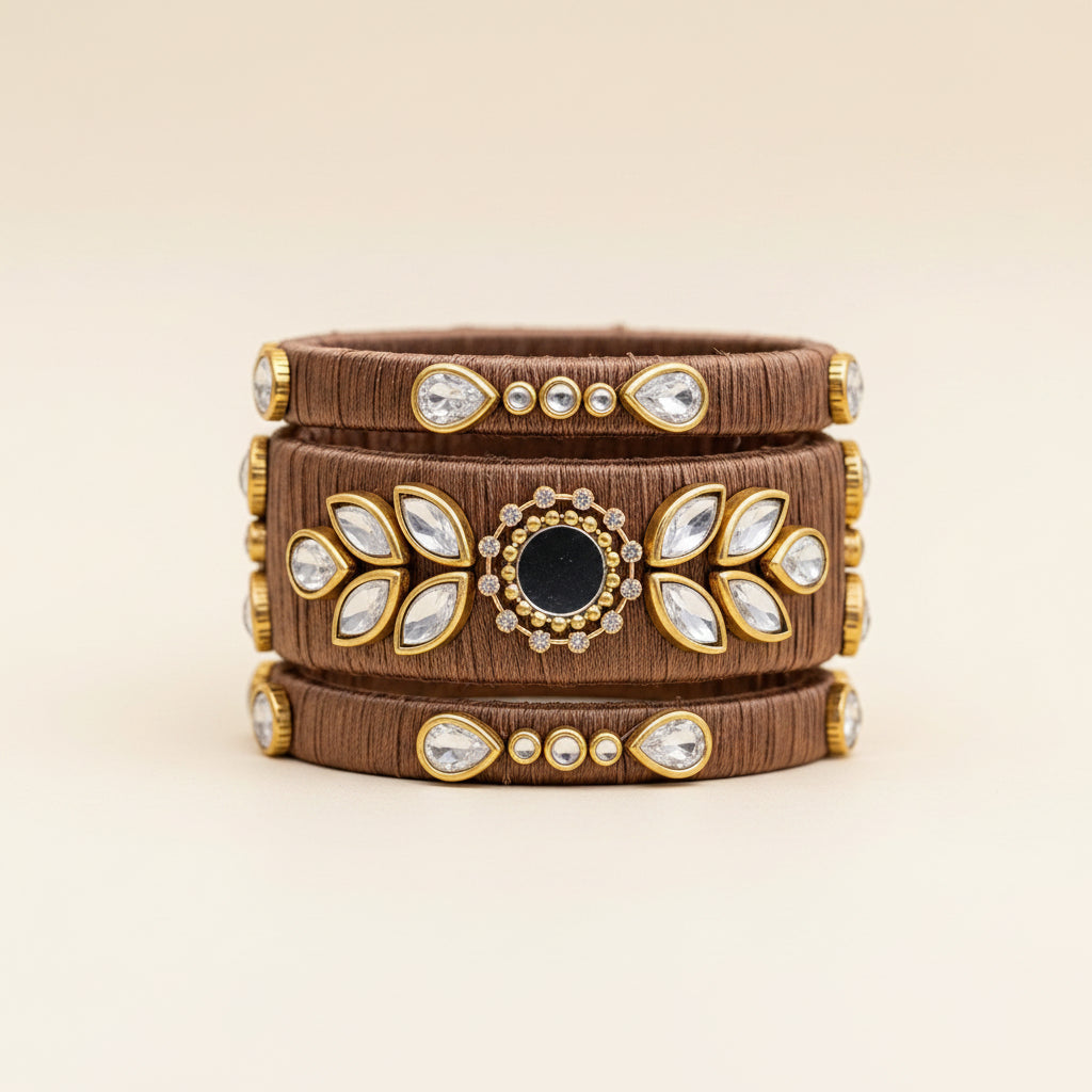 A wide brown fabric cuff bracelet with gold-tone metal trim, clear rhinestone accents, and a central dark oval stone, styled on a burlap surface with dried grass and eucalyptus leaves.