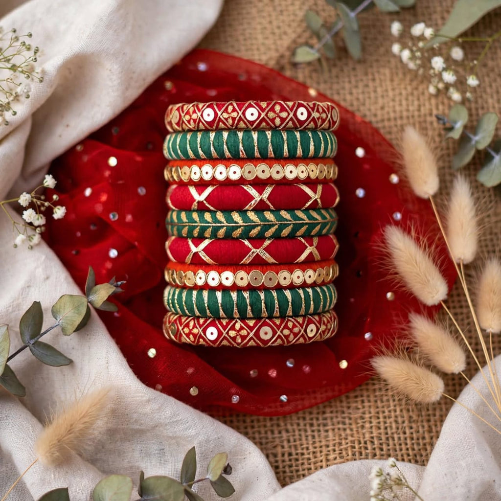 A stack of traditional Indian bangles with red, green, and gold embroidery and sequins, displayed on a red fabric cloth with decorative dried flowers and eucalyptus leaves.
