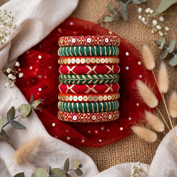 A stack of traditional Indian bangles with red, green, and gold embroidery and sequins, displayed on a red fabric cloth with decorative dried flowers and eucalyptus leaves.