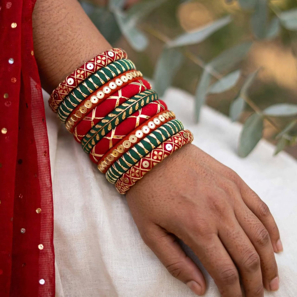 A close-up of a person's wrist and hand wearing a stack of traditional Indian bangles. The bangles are primarily red, green, and gold with intricate patterns and mirror work, worn over a white fabric and a red embroidered garment.