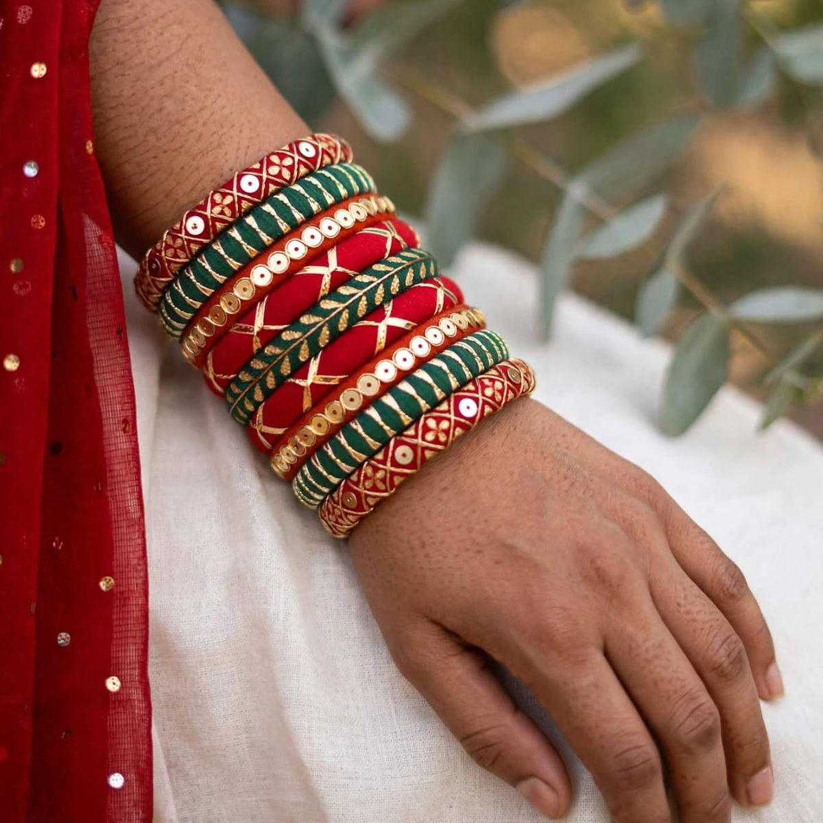 A close-up of a person's wrist and hand wearing a stack of traditional Indian bangles. The bangles are primarily red, green, and gold with intricate patterns and mirror work, worn over a white fabric and a red embroidered garment.