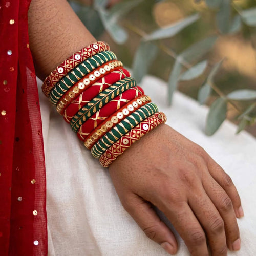 A close-up of a person's wrist and hand wearing a stack of traditional Indian bangles. The bangles are primarily red, green, and gold with intricate patterns and mirror work, worn over a white fabric and a red embroidered garment.