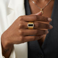 A close-up of a person's hand wearing a gold snake-print ring with a black rectangular center and a border of small clear stones, styled with a white blazer and dark gray vest.