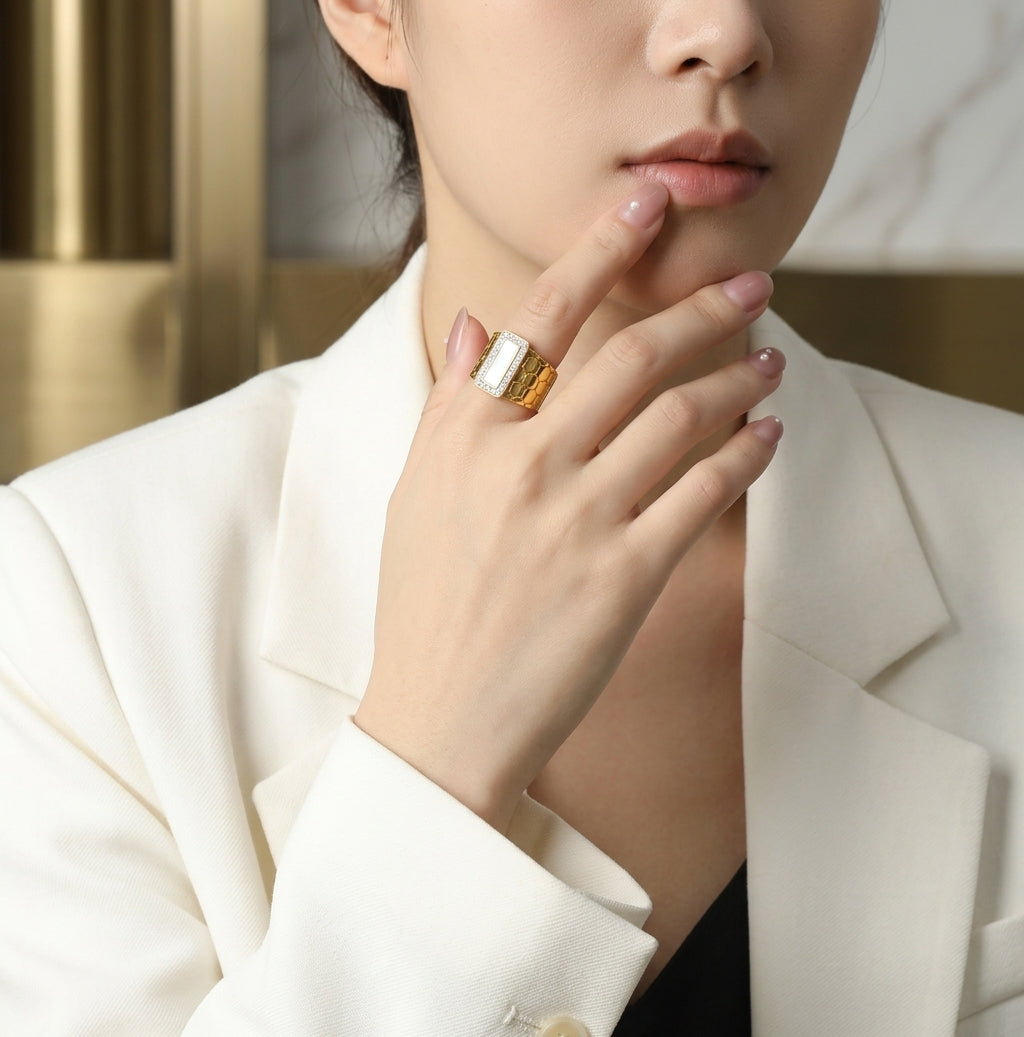A close-up of a woman wearing a large statement ring with a rectangular white stone and textured gold sides, styled with a white blazer.