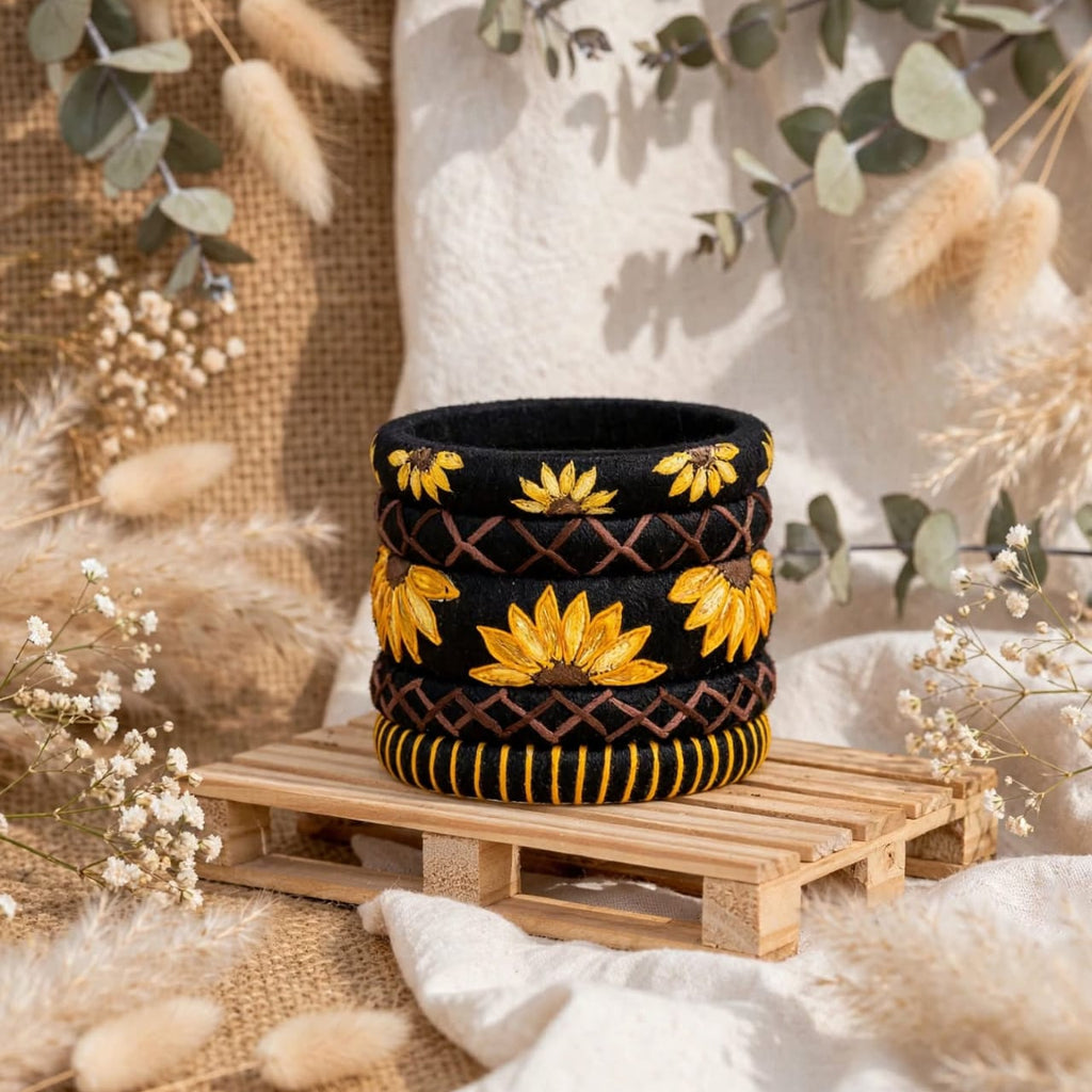 A black woven basket with yellow floral embroidery and brown geometric patterns, displayed on a small wooden pallet with neutral-toned dried flowers and greenery in the background.