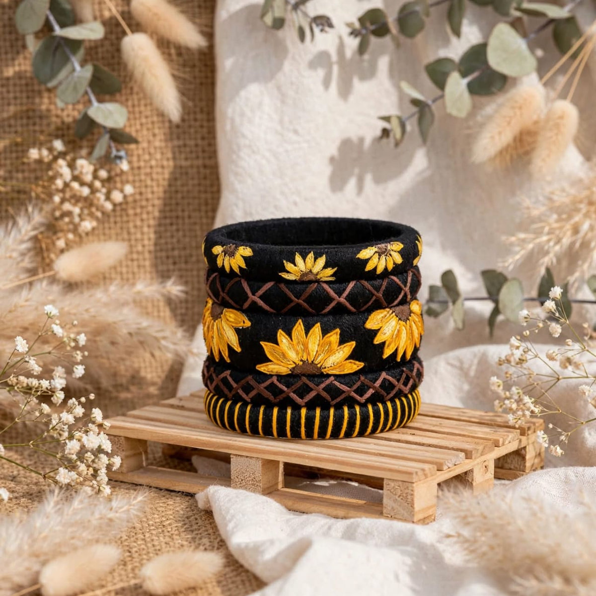 A black woven basket with yellow floral embroidery and brown geometric patterns, displayed on a small wooden pallet with neutral-toned dried flowers and greenery in the background.
