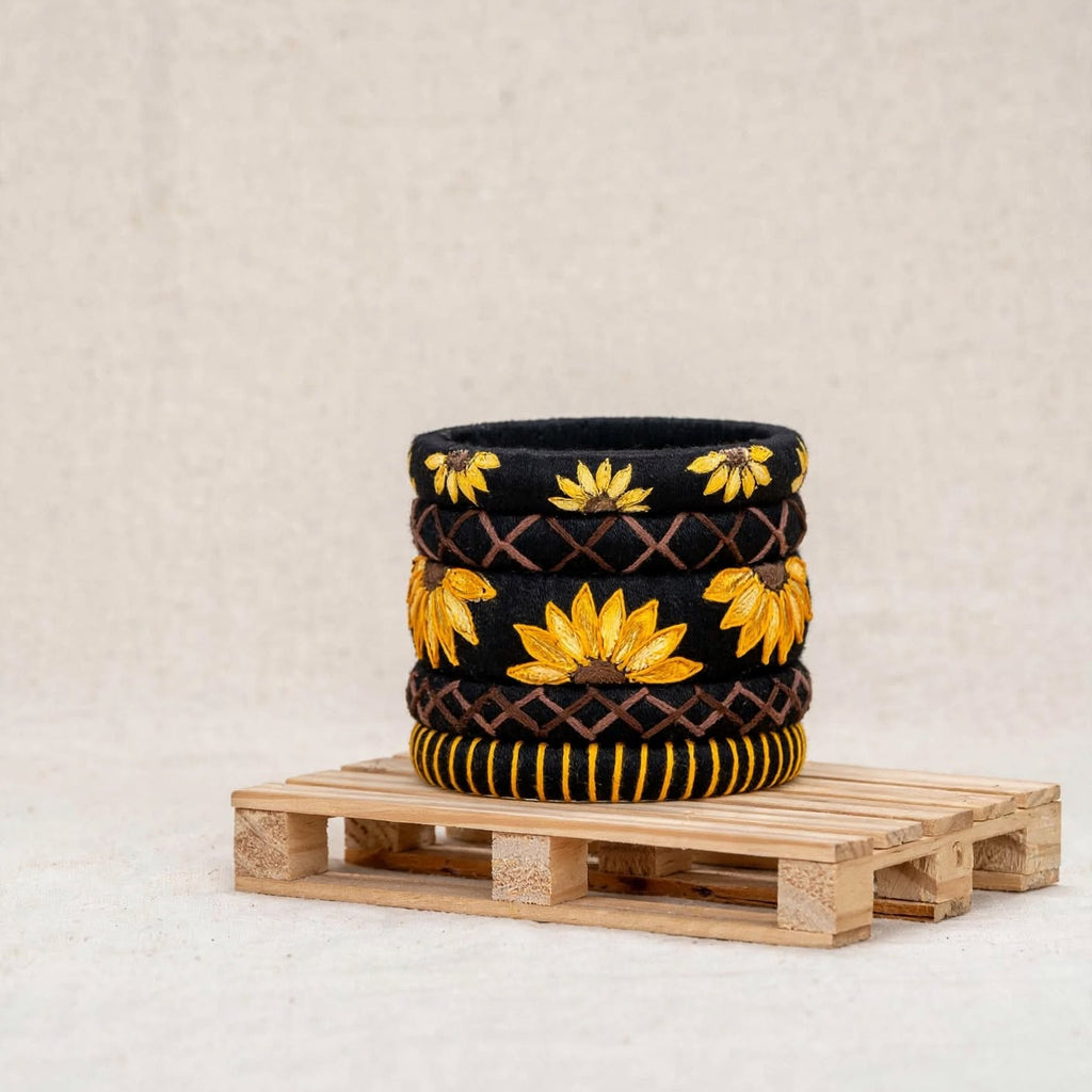 A black knit cowl with yellow sunflower and brown argyle patterns, displayed on a small wooden pallet against a neutral beige background.
