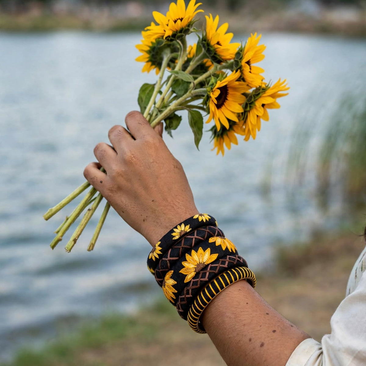 A person's hand holding a bouquet of yellow sunflowers near a body of water, wearing a black and yellow patterned woven cuff bracelet.