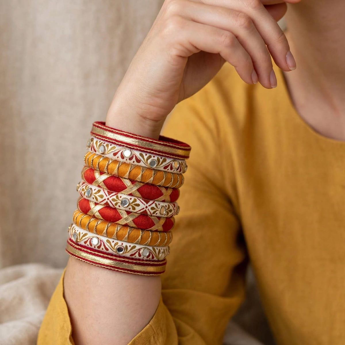 A close-up of a person wearing a stack of traditional Indian bangles on their wrist. The bangles feature intricate gold, red, and white patterns, including a central red and gold geometric design.