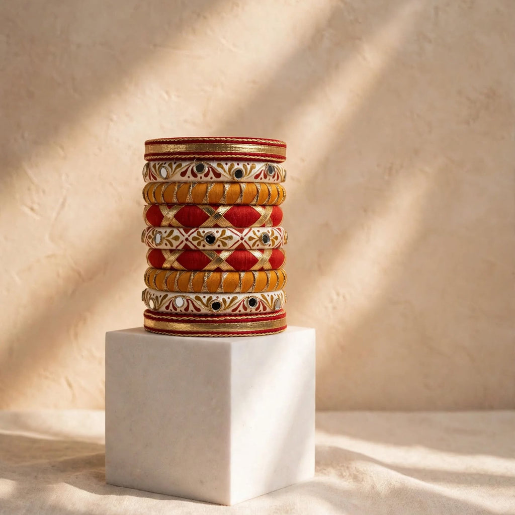 A stack of ornate traditional Indian bangles with red, gold, and white patterns, displayed on a white cube pedestal against a textured beige wall with warm sunlight shadows.