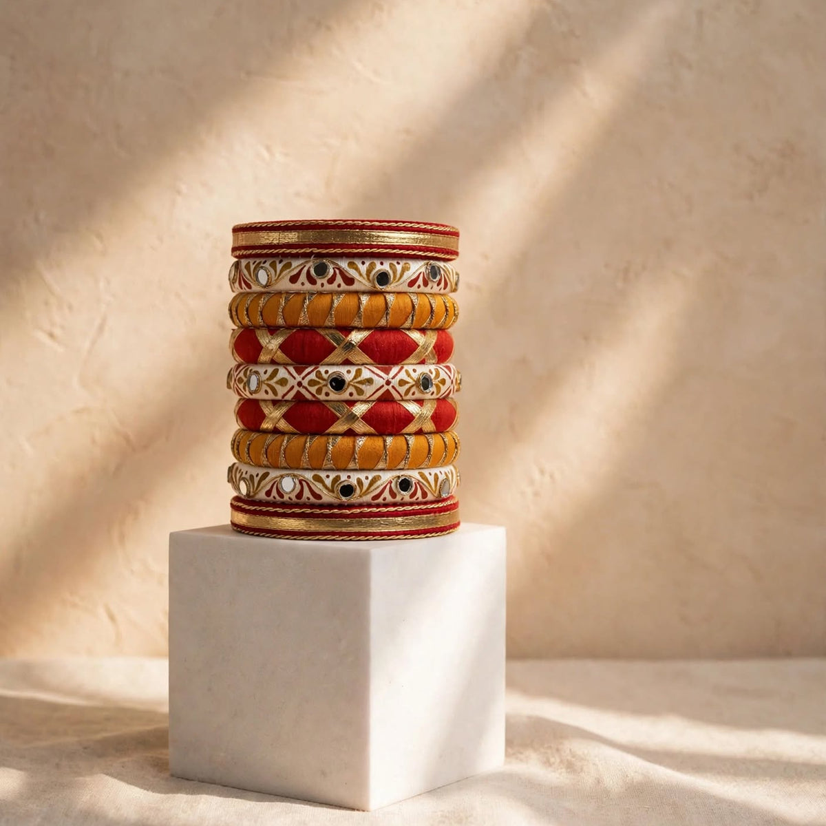 A stack of ornate traditional Indian bangles with red, gold, and white patterns, displayed on a white cube pedestal against a textured beige wall with warm sunlight shadows.