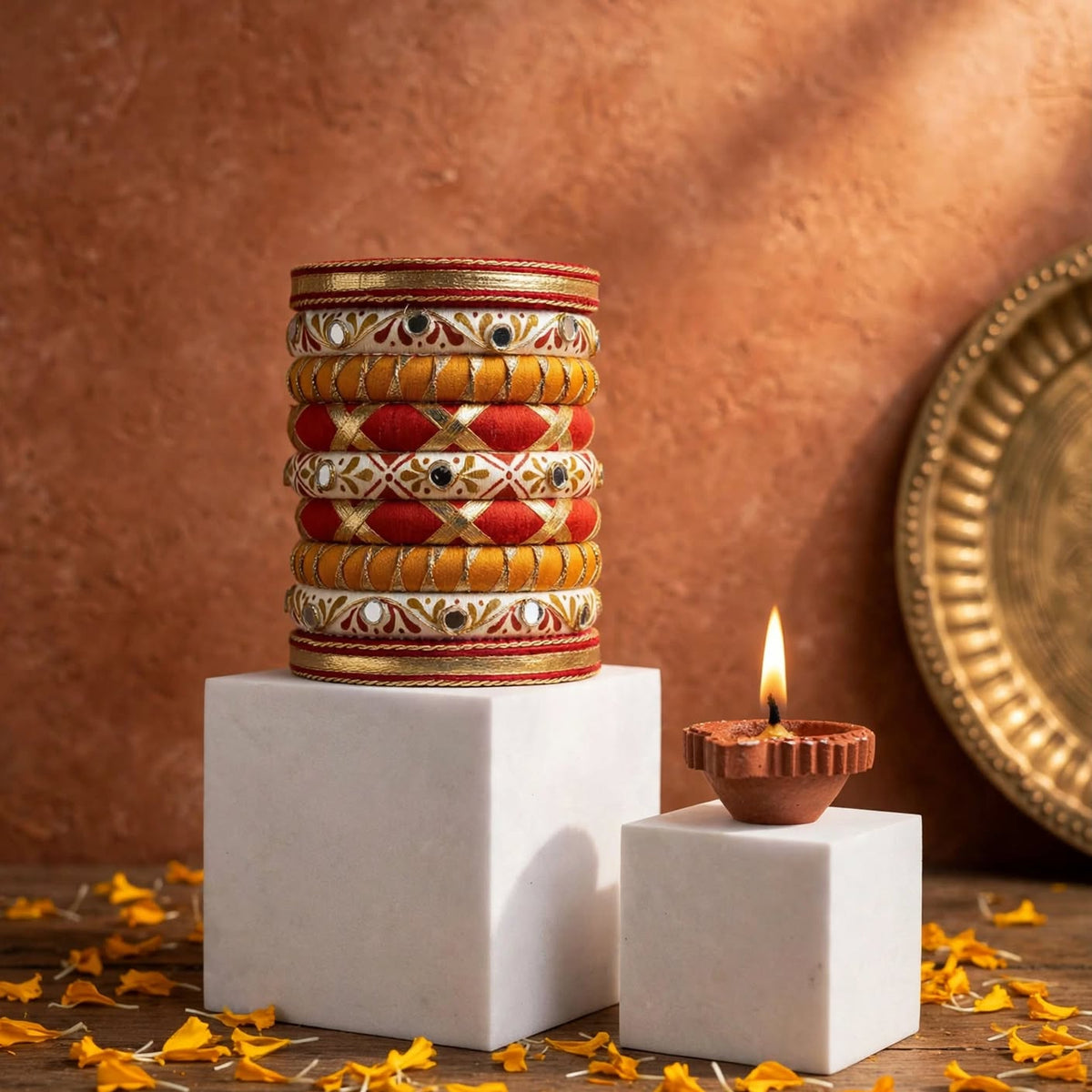 A stack of ornate Indian bangles with red, gold, and white patterns displayed on white pedestals next to a lit clay diya, with scattered orange petals on a wooden surface against a textured terracotta background.
