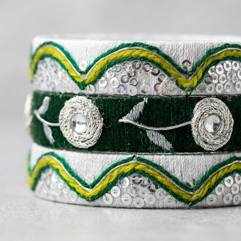 A close-up of a traditional Indian silver-toned bangle bracelet featuring a dark green central band with silver floral motifs, bordered by wavy green and yellow embroidered trim and silver sequins on a light gray background.