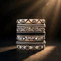 A stack of five patterned ceramic coasters with black geometric and floral designs on a cream-colored glaze, photographed under dramatic spotlighting on a dark background.