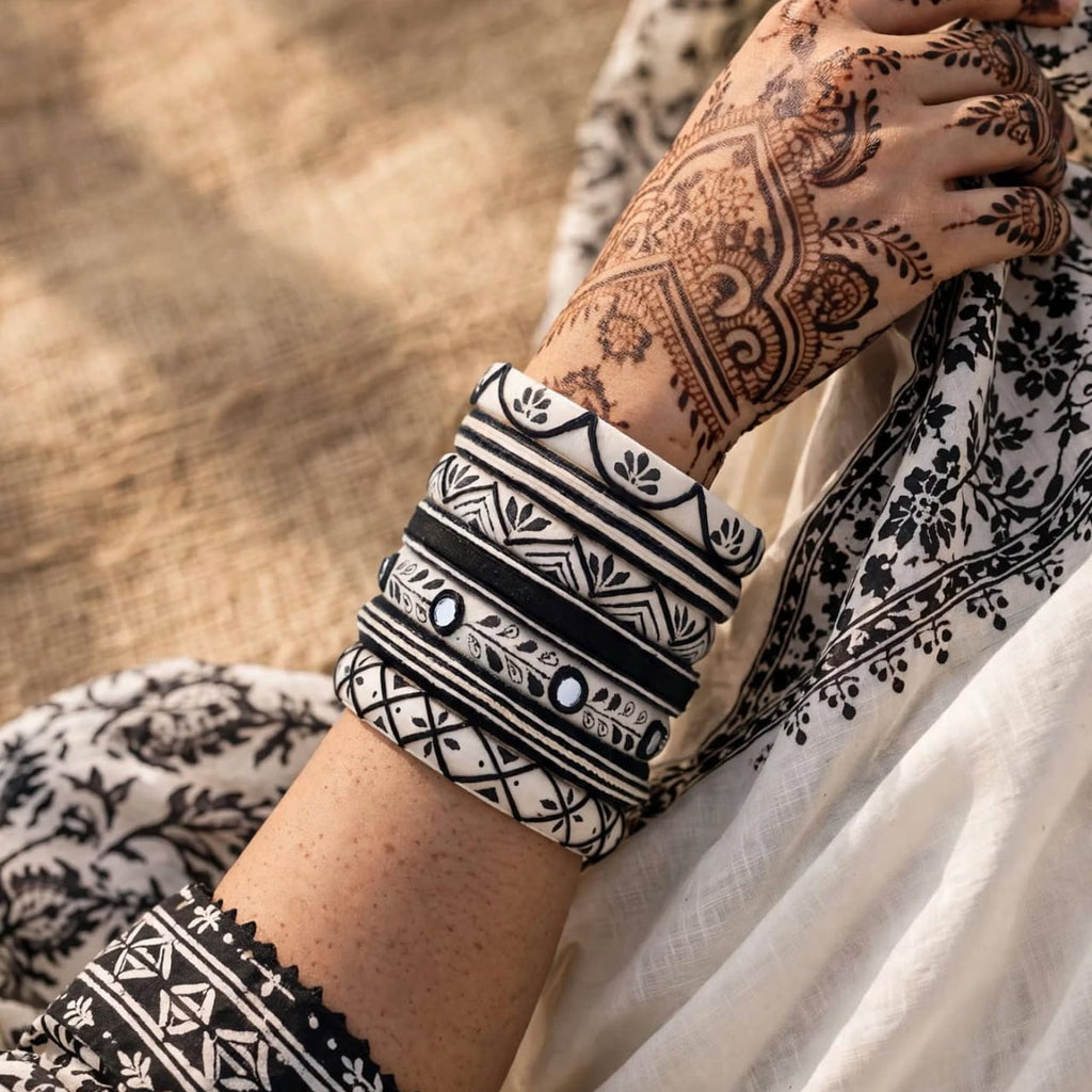 A close-up of a person's hand with intricate brown henna, wearing a stack of wide black and white patterned bangles, resting on a white and black patterned fabric against a textured beige background.
