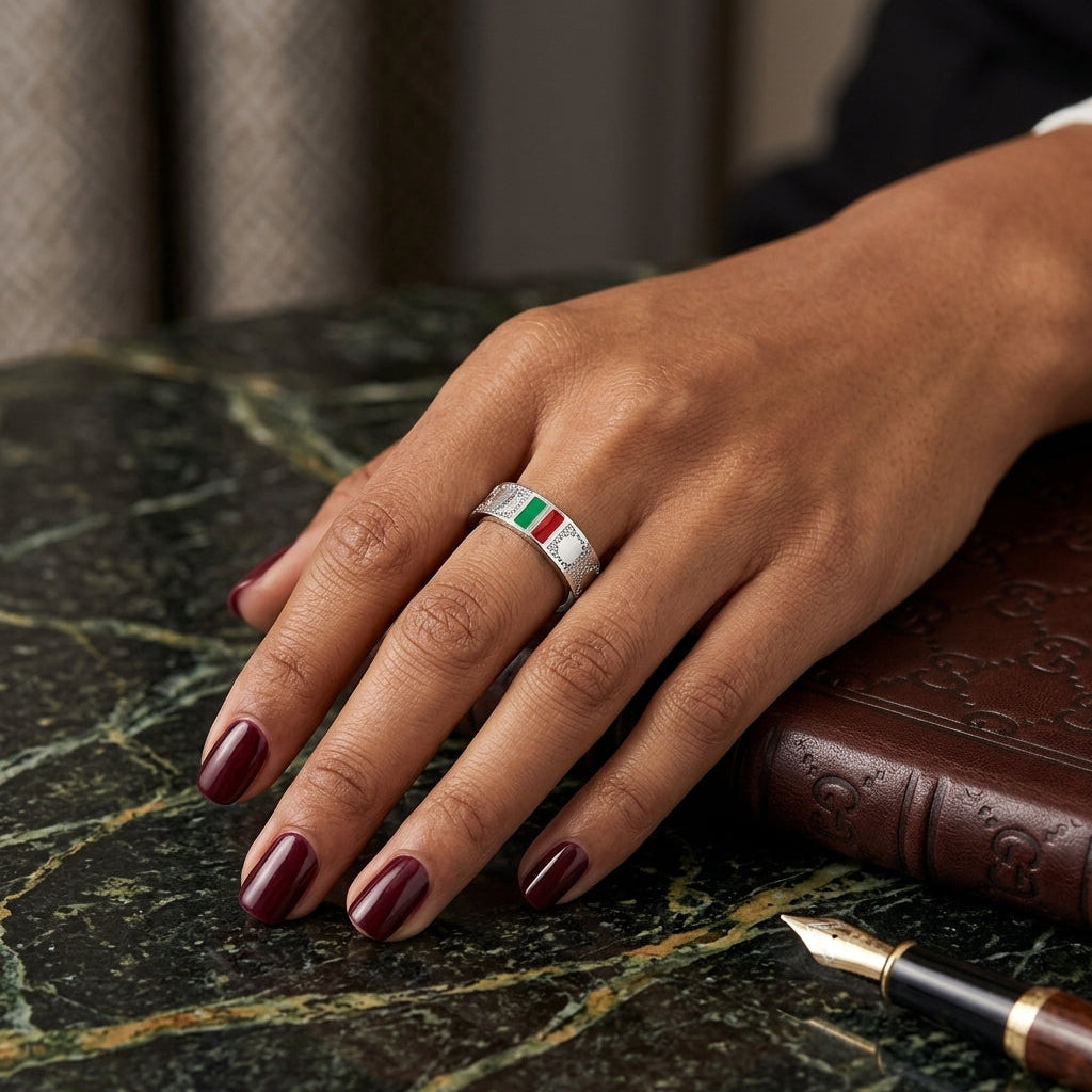 A silver ring with a central green and white striped inlay, worn on a hand with dark burgundy nail polish, resting on a green marble surface next to a brown leather-bound book.