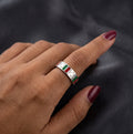 A close-up of a hand wearing a silver ring with a central rectangular inlay featuring green, white, and red stripes, set against a dark textured background.