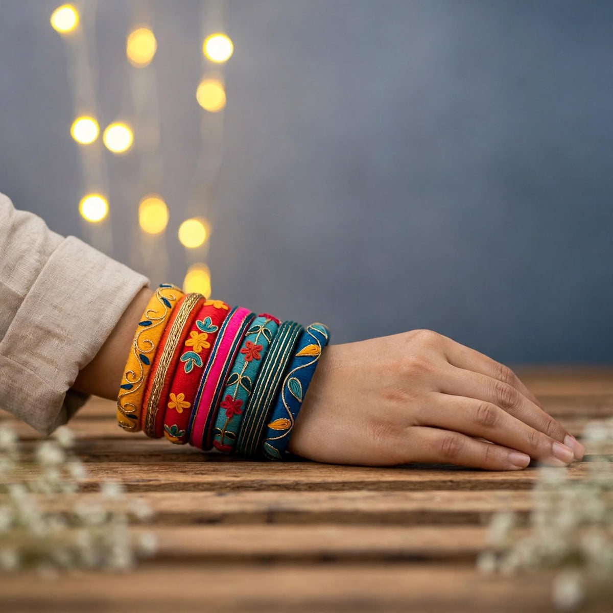 A close-up of a person's hand resting on a wooden surface, adorned with a stack of colorful embroidered bangles. The background features a soft, out-of-focus blue wall with warm bokeh string lights.