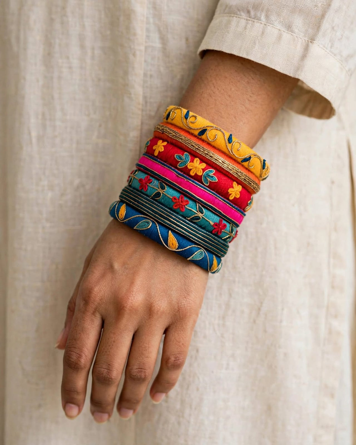 A close-up of a person's wrist wearing a stack of colorful embroidered bangles (kara bracelets) in yellow, red, pink, blue, and green, paired with a light beige linen kurta.