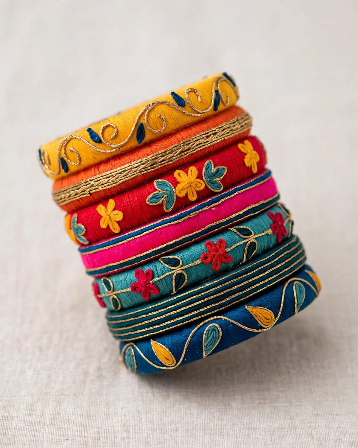 A stack of six colorful embroidered bangles on a light beige textured fabric background. The bangles feature vibrant colors including yellow, orange, red, pink, teal, and blue, with detailed floral and vine embroidery.