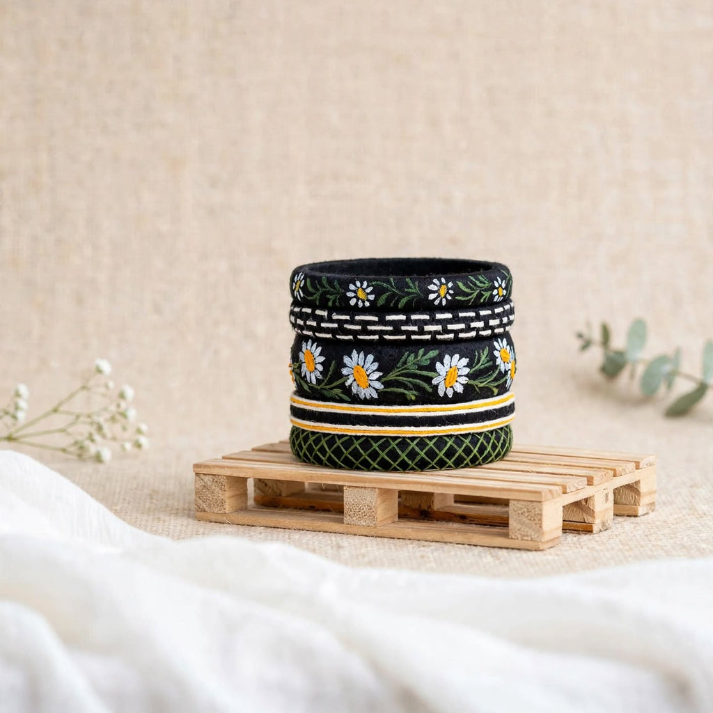 A black ceramic cup with a white daisy and green leaf pattern, displayed on a small wooden stand against a neutral beige background.