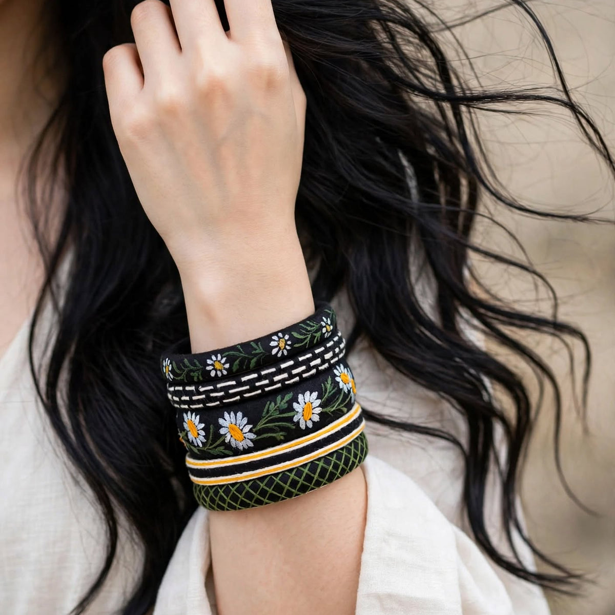 A close-up of a person's wrist wearing a wide black cuff bracelet decorated with embroidered white daisies, yellow centers, and green stems, along with white and yellow stripes.
