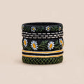 A cylindrical woven basket or planter with a black base, decorated with embroidered white daisies, green leaves, and geometric patterns, set against a solid beige background.