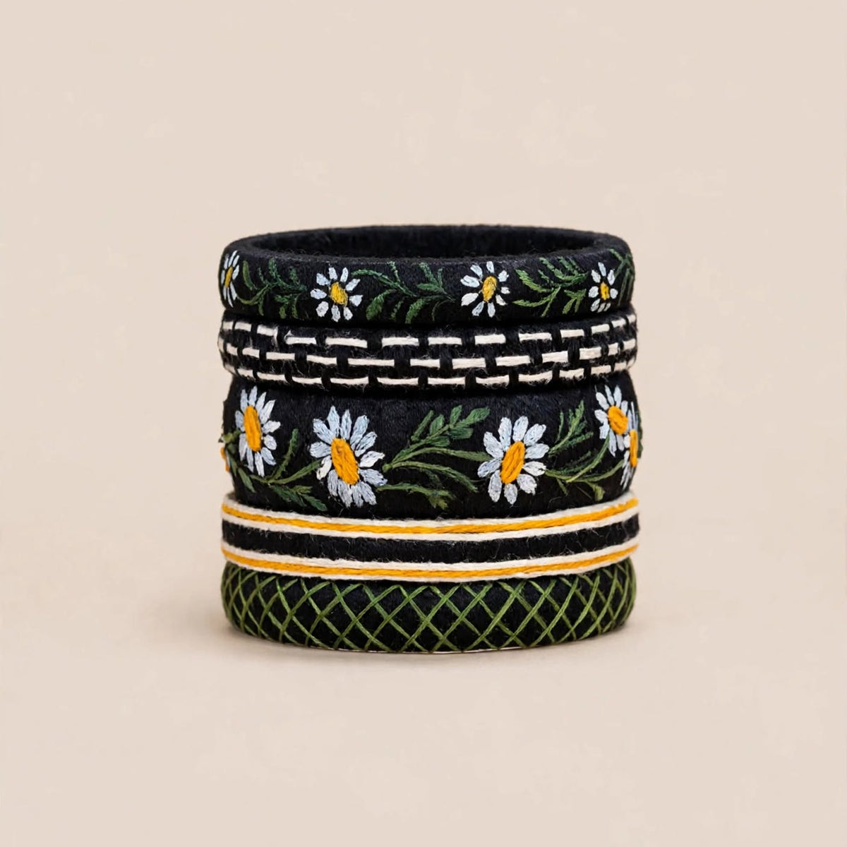 A cylindrical woven basket or planter with a black base, decorated with embroidered white daisies, green leaves, and geometric patterns, set against a solid beige background.