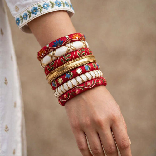 A close-up of a person's wrist and hand wearing a stack of traditional Indian bangles, including red, white, and gold embroidered styles, against a neutral beige background.