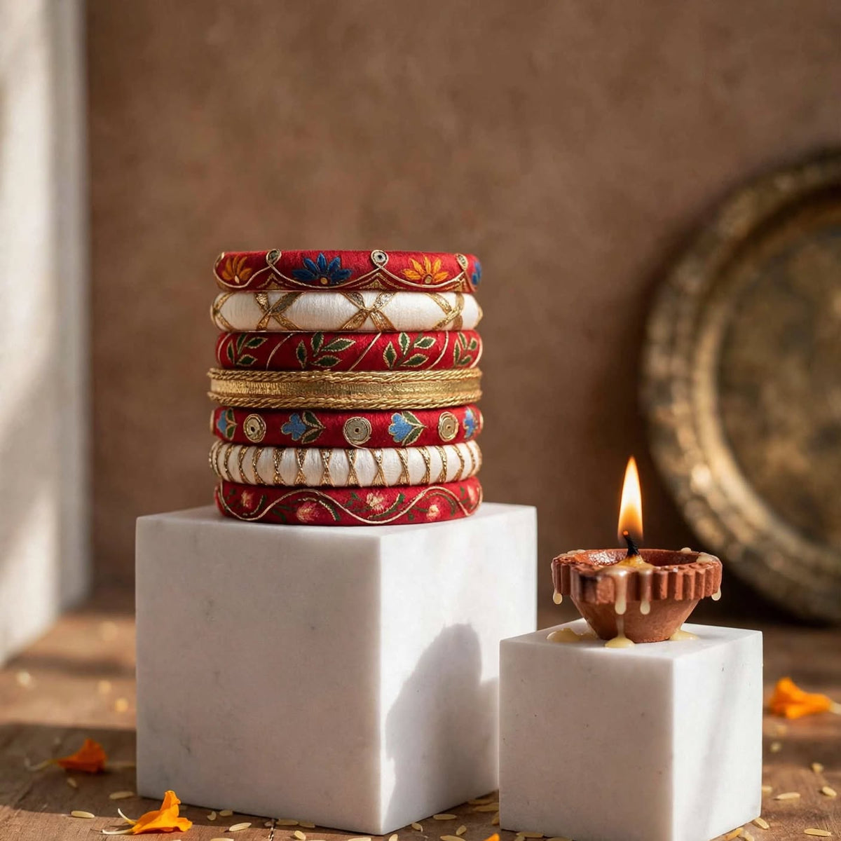 A stack of colorful traditional Indian bangles displayed on white marble pedestals, with a small lit diya candle on a matching pedestal against a rustic brown background.