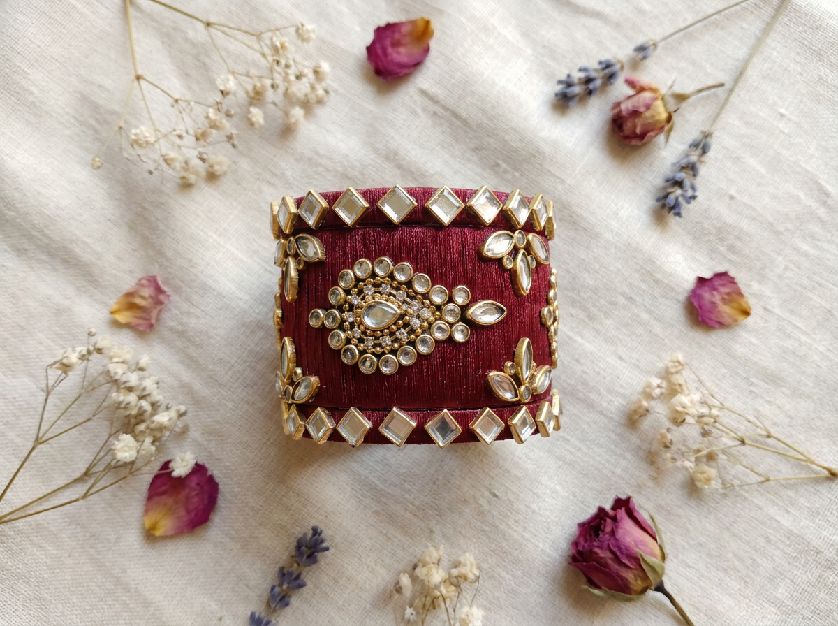 A cylindrical deep red velvet cuff bracelet decorated with gold-toned metallic borders, rhinestones, and a central floral embellishment, styled on a textured cream fabric with dried flowers.