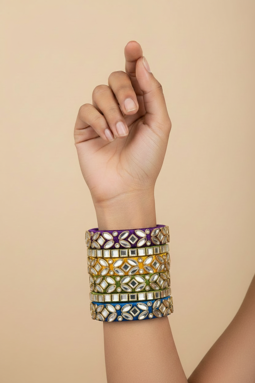 A close-up of a person's hand and wrist wearing a stack of five colorful bangles with mirror work and colored borders, set against a solid beige background.