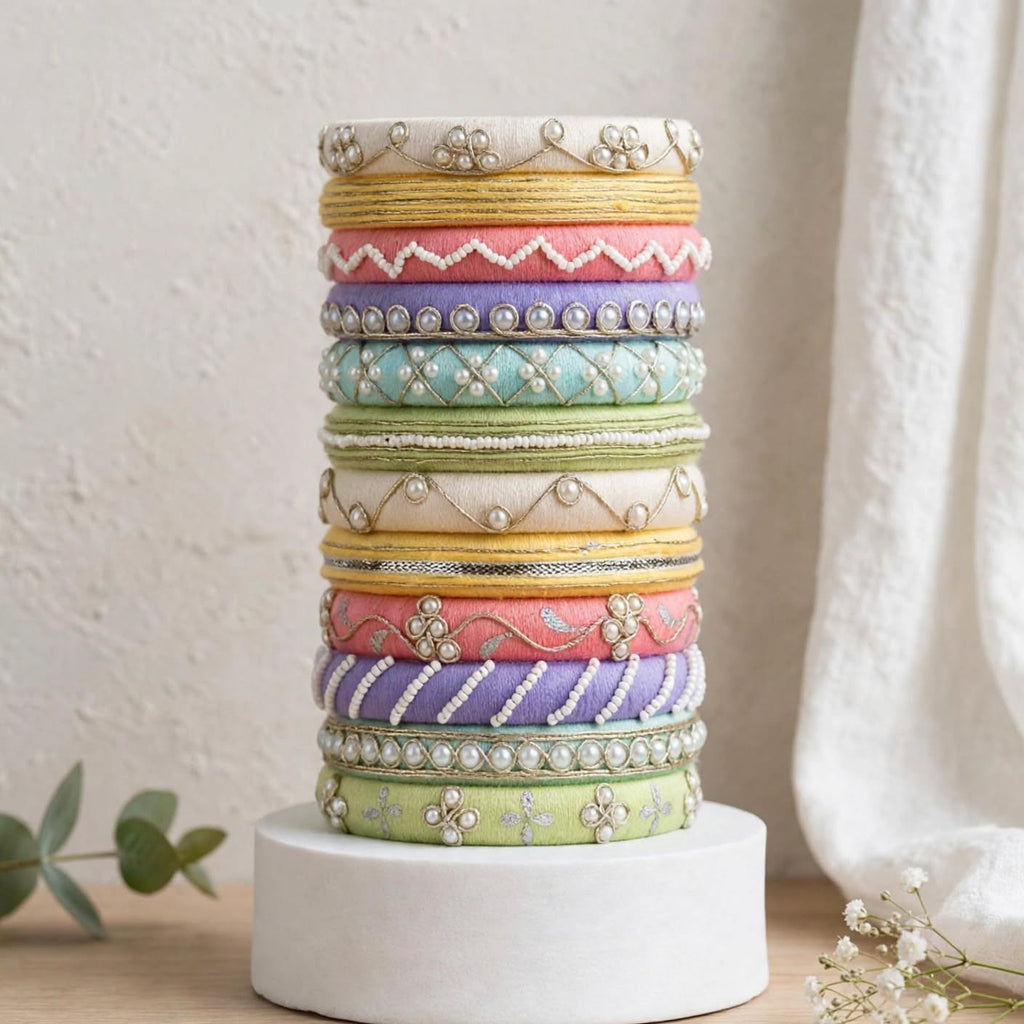 A stack of colorful embroidered bangles displayed on a white cylindrical pedestal. The bangles feature various patterns including floral, geometric, and beadwork in pastel shades of yellow, pink, purple, blue, green, and white, set against a soft, neutral background with a textured wall and draped fabric.