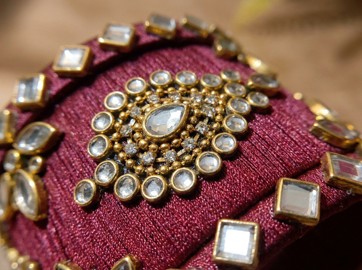 A close-up of an ornate gold-toned jewelry piece with a central teardrop-shaped clear stone surrounded by round and square clear stones, set on a deep burgundy embroidered fabric background.