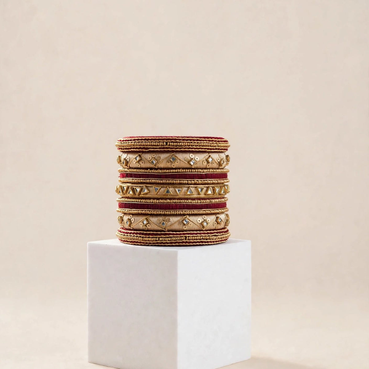 A stack of ornate gold-toned bangles with red and gold embroidered borders and small clear stone accents, displayed on a white cube against a warm beige background.