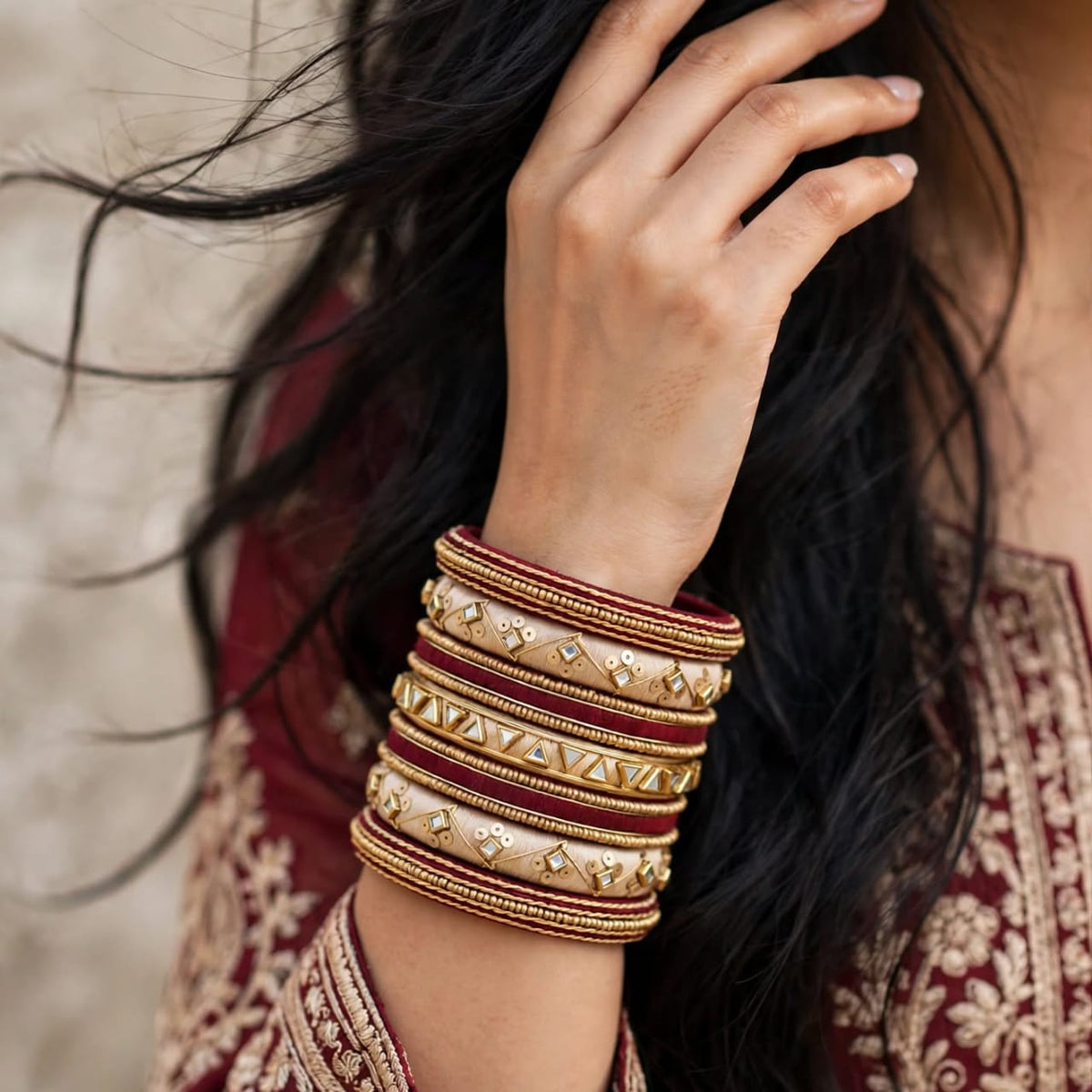 A close-up of a person wearing a stack of traditional Indian bangles, featuring gold-plated cuffs with red inlay and small diamond-shaped embellishments, paired with a matching red and gold embroidered outfit.