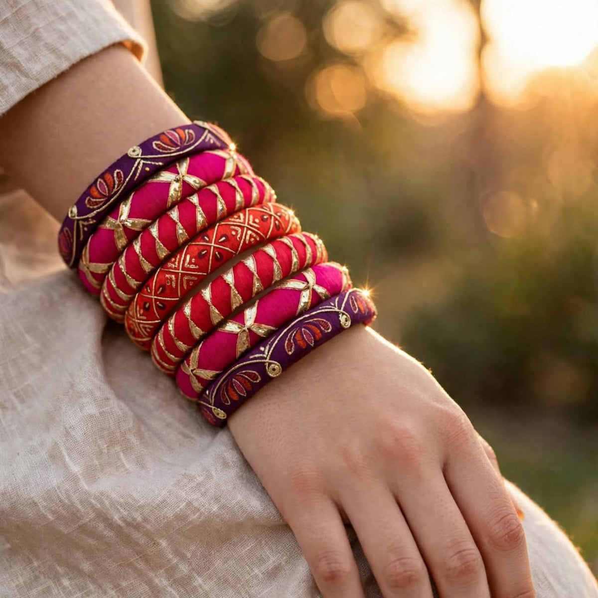 A close-up of a person's wrist wearing a stack of ornate, colorful embroidered bangles. The bangles feature intricate patterns in pink, red, purple, and gold, set against a soft-focus outdoor background with warm sunlight.
