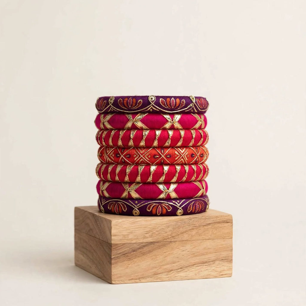 A stack of eight traditional Indian bangles (kara bracelets) in vibrant shades of magenta, orange, and deep purple, decorated with gold foil work and red floral patterns, displayed on a wooden block against a warm off-white background.