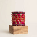 A stack of eight traditional Indian bangles (kara bracelets) in vibrant shades of magenta, orange, and deep purple, decorated with gold foil work and red floral patterns, displayed on a wooden block against a warm off-white background.