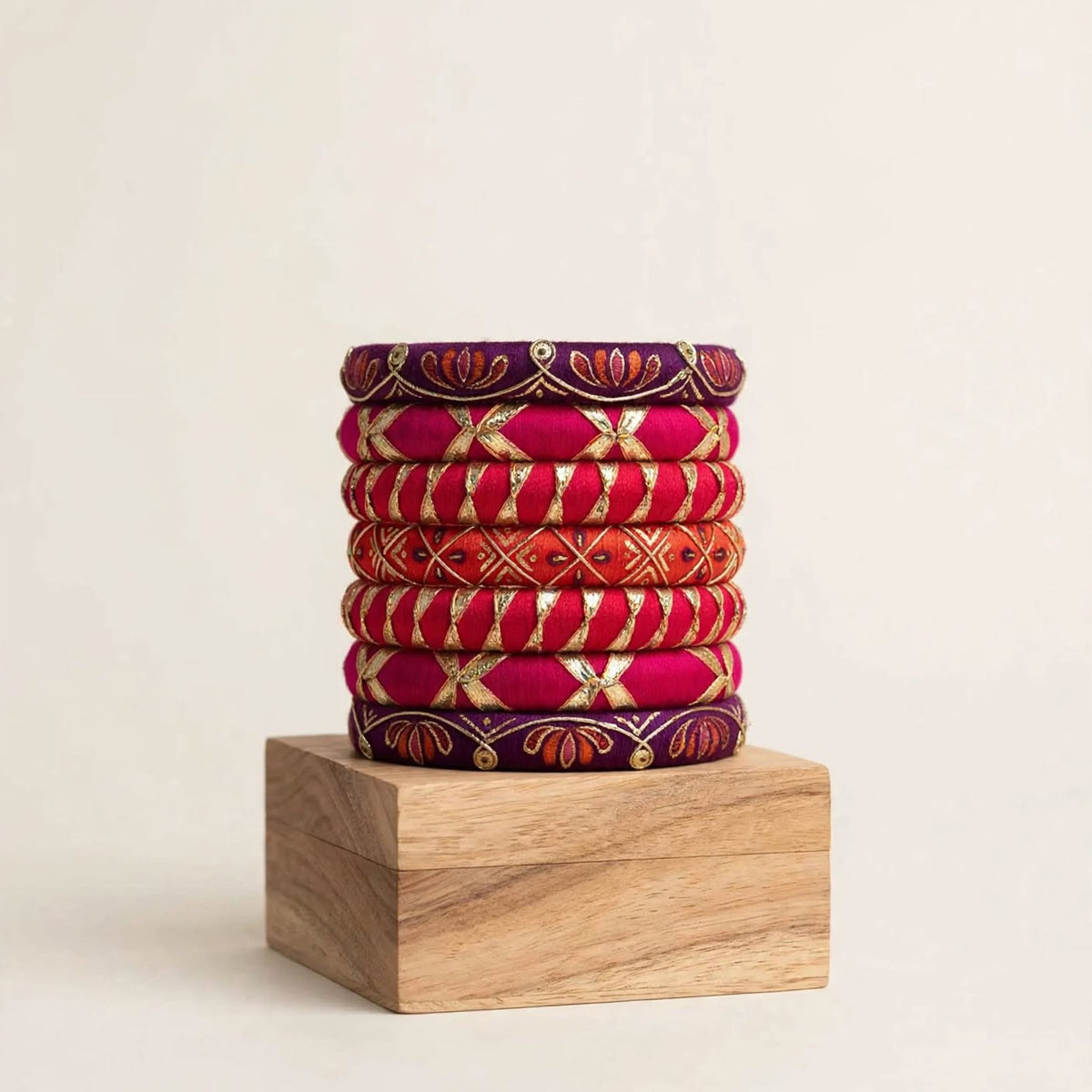 A stack of eight traditional Indian bangles (kara bracelets) in vibrant shades of magenta, orange, and deep purple, decorated with gold foil work and red floral patterns, displayed on a wooden block against a warm off-white background.