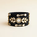 A stack of three black elastic bangles decorated with gold-toned metal frames, white stones, and a central round white stone, displayed on a draped light purple fabric background.