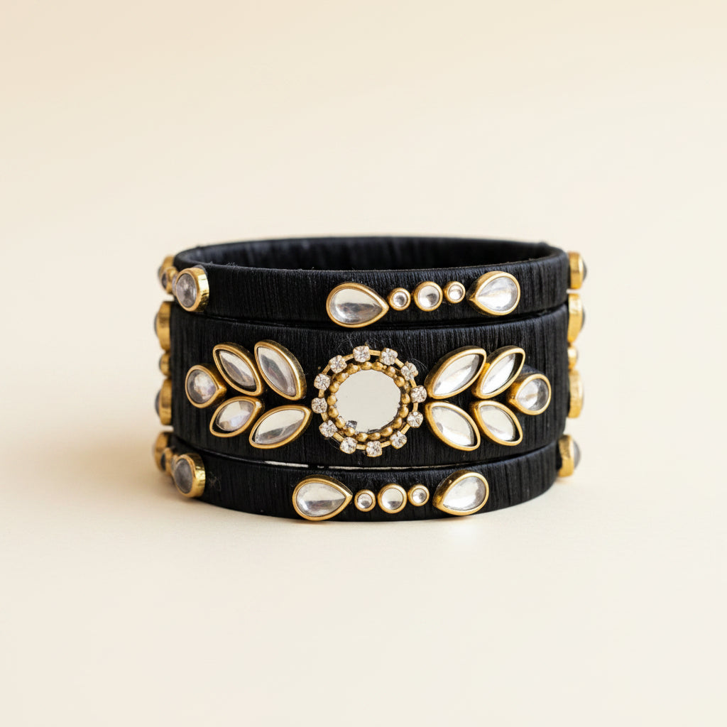 A stack of three black elastic bangles decorated with gold-toned metal frames, white stones, and a central round white stone, displayed on a draped light purple fabric background.