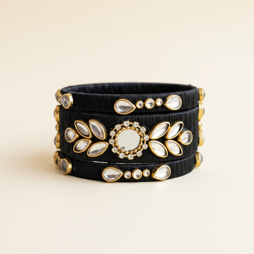 A stack of three black elastic bangles decorated with gold-toned metal frames, white stones, and a central round white stone, displayed on a draped light purple fabric background.