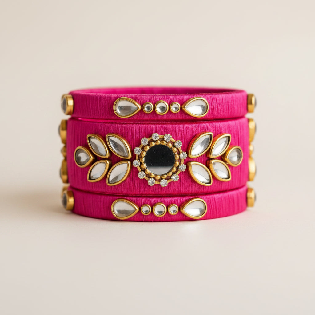 A bright pink fabric cuff bracelet with gold-toned metal accents, white mirror-like stones, and a central black stone, styled on a beige textured surface with warm fairy lights and dried grass.