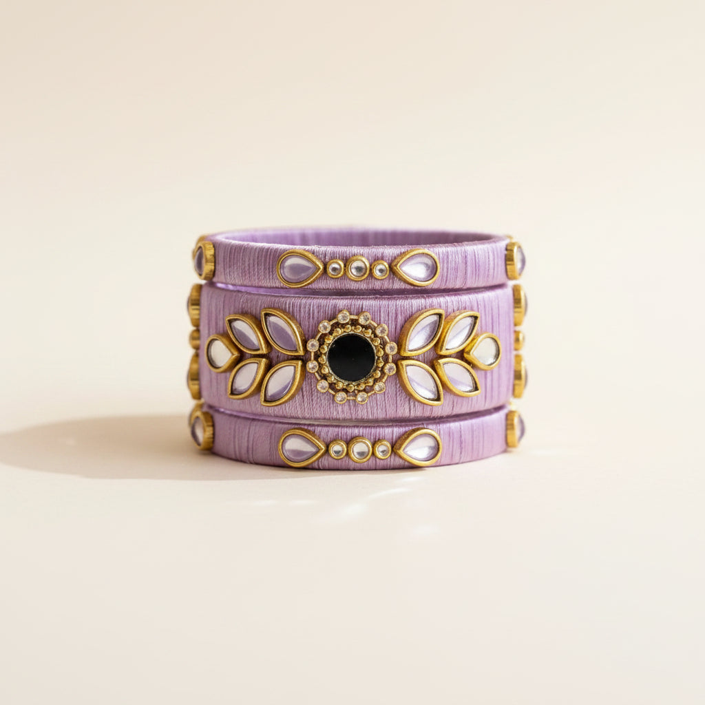 A lavender wrap bracelet with gold-toned filigree and a central black stone, styled on a textured lavender fabric with dried grass and baby's breath flowers.