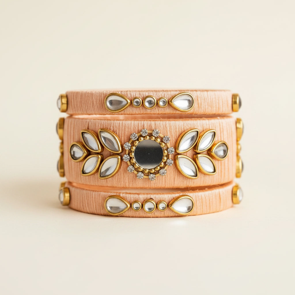 A wide three-band cuff bracelet in peach-colored textured leather with gold-tone metal accents, clear teardrop rhinestones, and a central round dark stone, styled on a peach fabric background with dried grasses and eucalyptus.