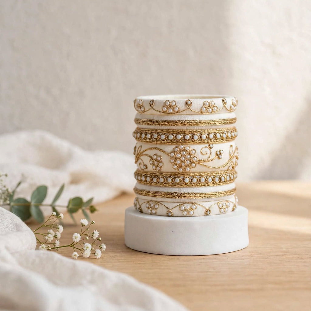A stack of five white cylindrical candle jars with ornate gold floral and vine patterns, displayed on a white circular base on a wooden surface with soft white fabric and small flowers in the foreground.